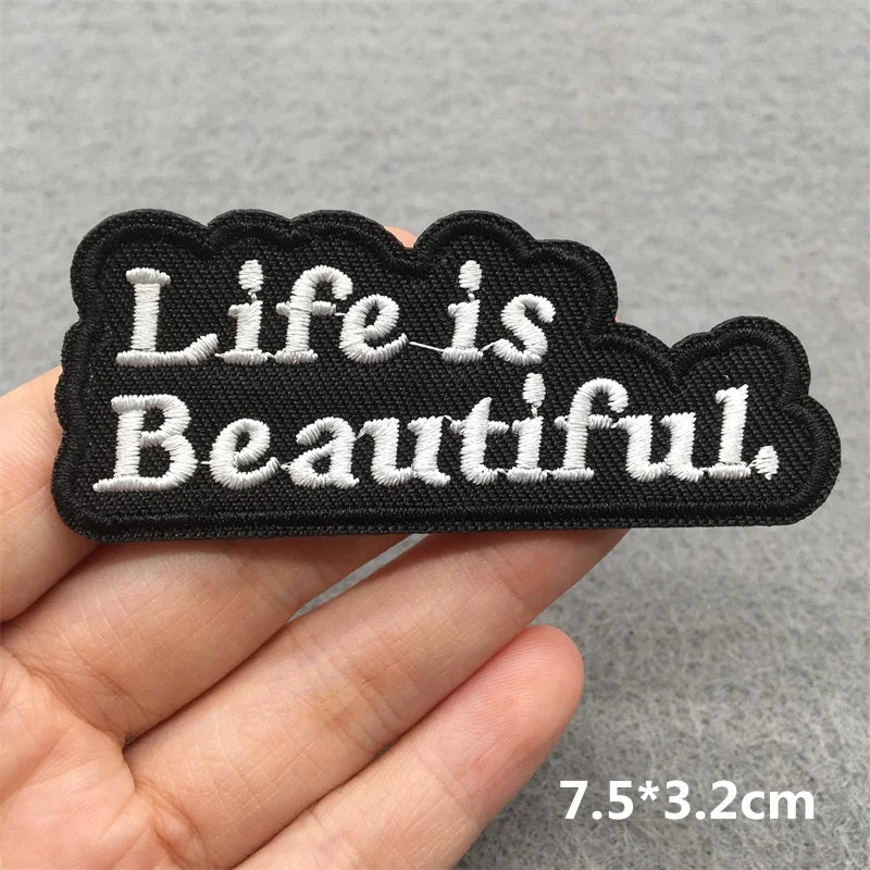 Embroidered patch with 'Life is Beautiful' text held by a hand on a gray background