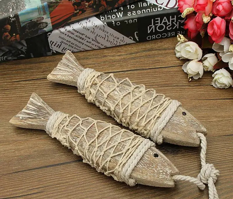 Two wooden fish with rope on a wooden surface with flowers and books in the background.