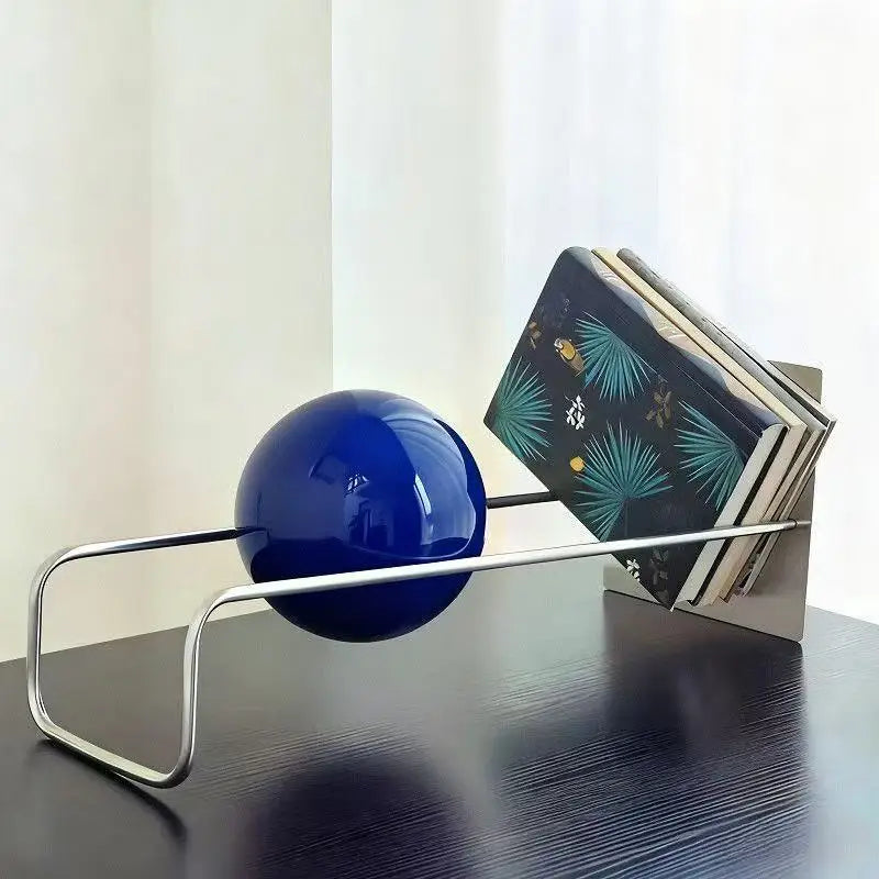 A modern metal bookend with a glossy dark blue spherical weight, shown holding a stack of books on a dark wooden table against a light curtain background.