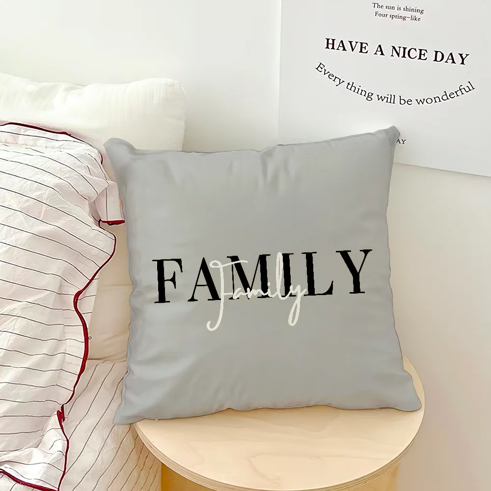 A square light gray throw pillow with the word 'FAMILY' printed in black and white script, styled on a round wooden side table next to a striped pillow against a white wall with a decorative print.