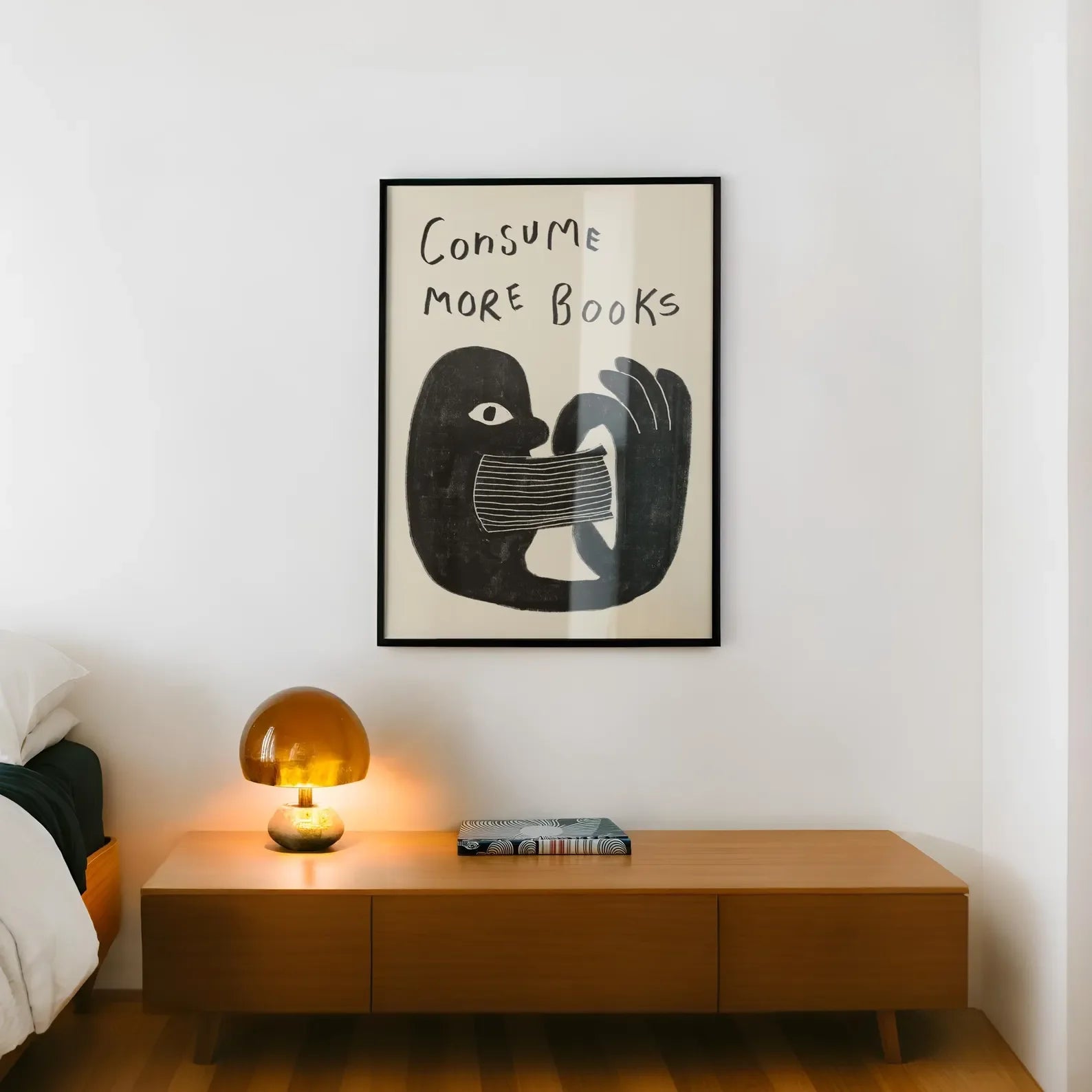 Framed artwork with a hand holding a book and 'Consume More Books' text above a wooden nightstand with a lamp.