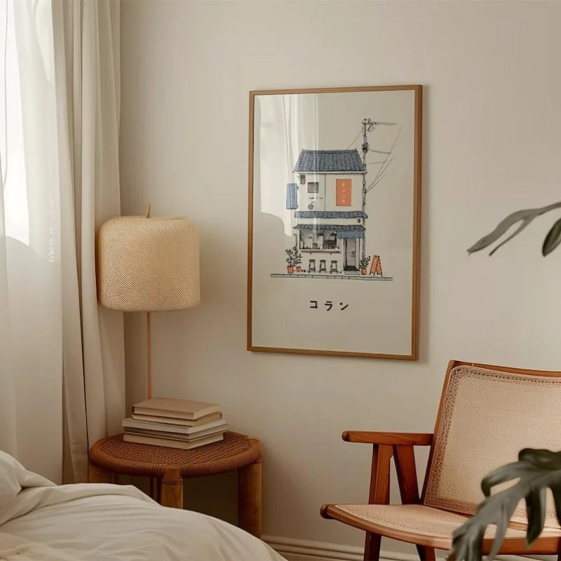 A framed art print depicting a small Japanese-style house with the word 'デード' below it, hanging on a beige wall in a cozy bedroom setting next to a lamp, side table, and wooden chair.
