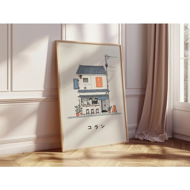 A framed art print featuring a stylized illustration of a Japanese street shop with the word 'Coran' in Japanese characters, leaning against a paneled wall on a wooden floor next to a window with curtains.