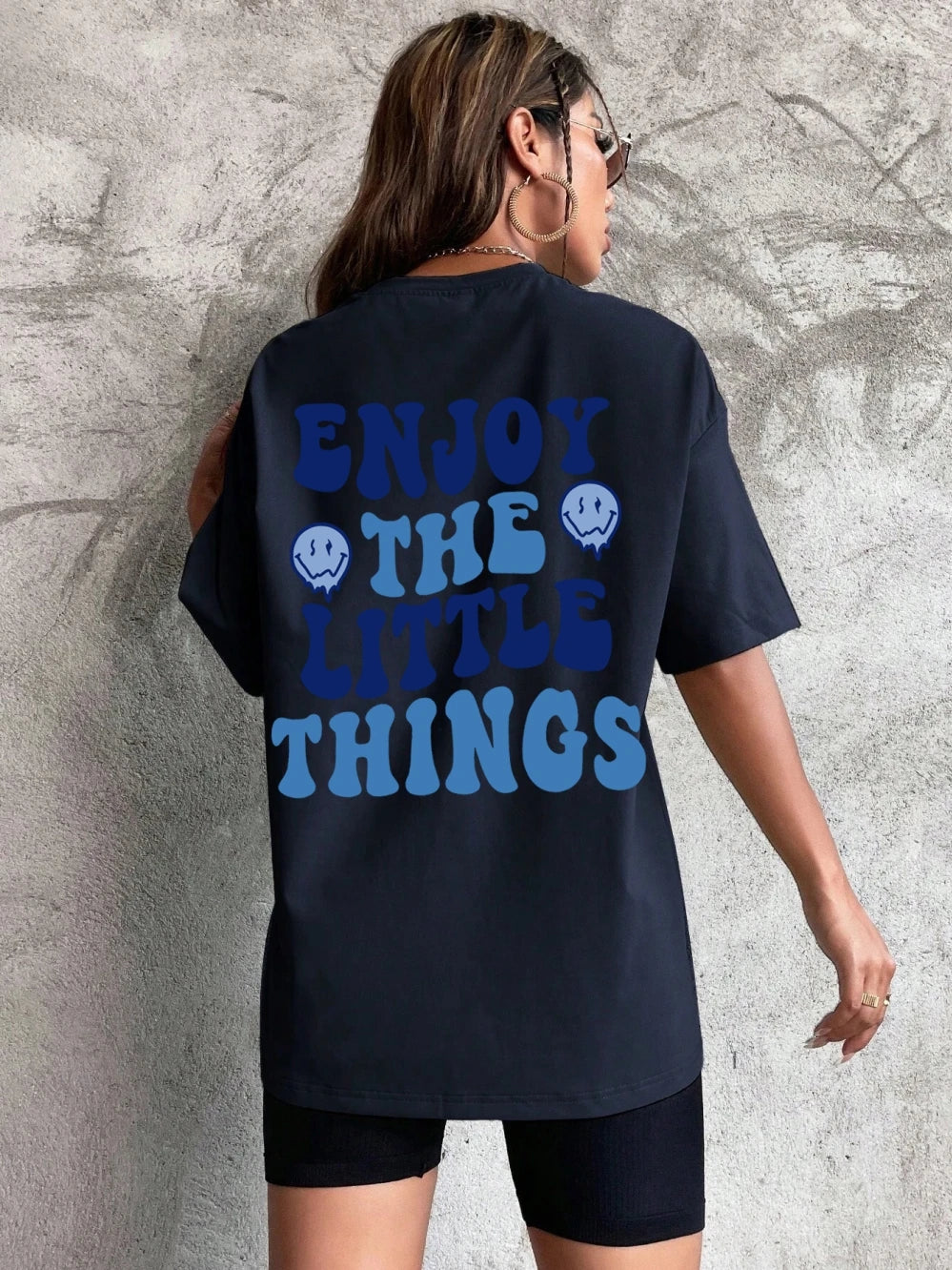 Person wearing a black t-shirt with 'Enjoy the Little Things' text on the back against a gray concrete wall.
