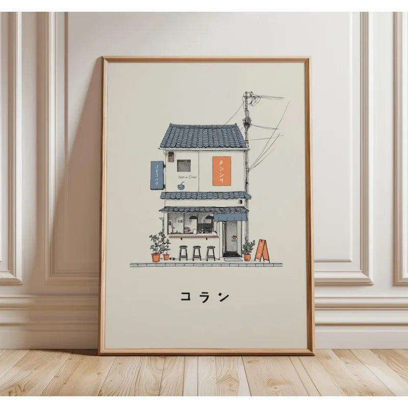 A framed art print featuring a minimalist illustration of a Japanese street cafe with Japanese text, leaning against a cream-colored paneled wall on a light wood floor.