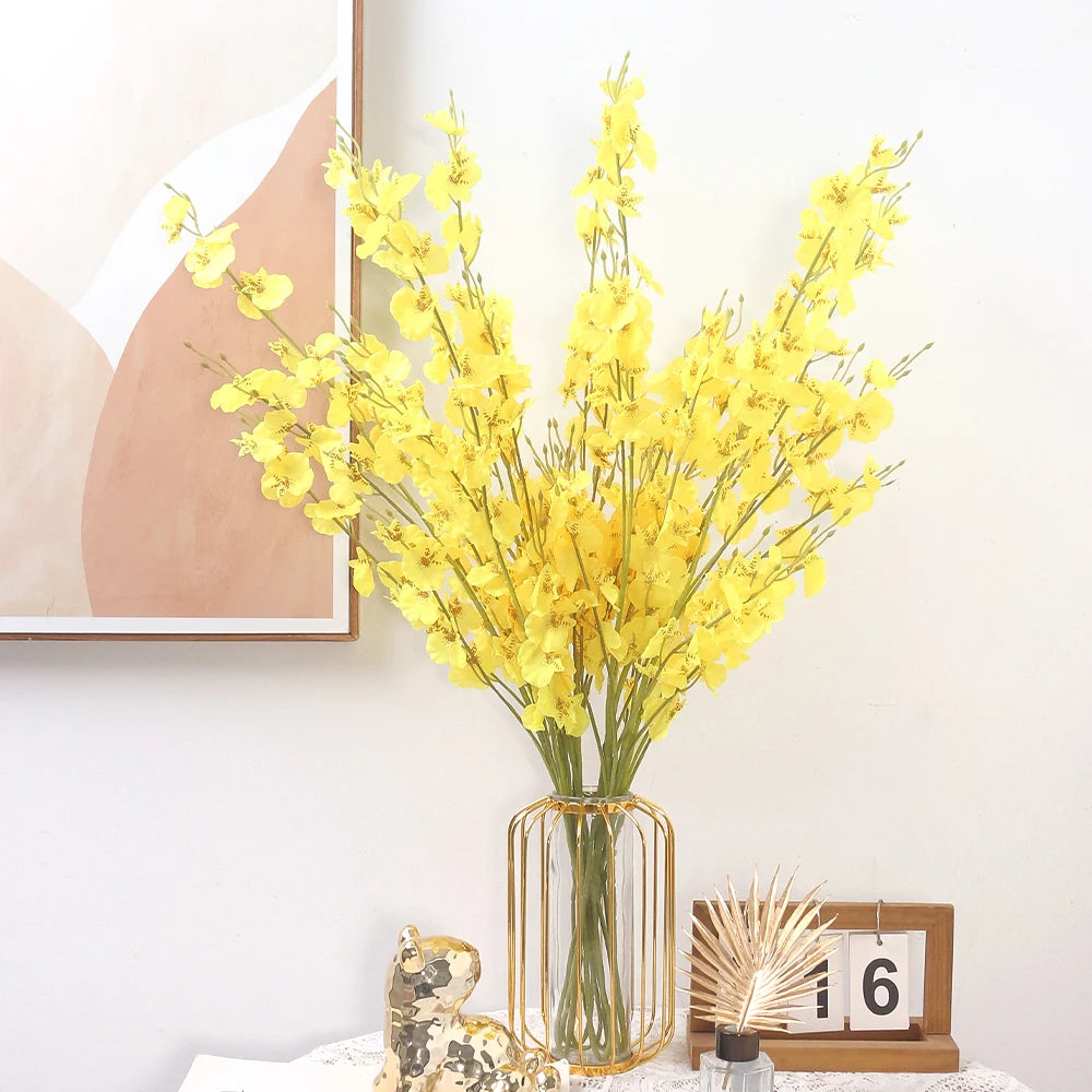 Yellow flowers in a glass vase on a white surface with a framed abstract art piece in the background.