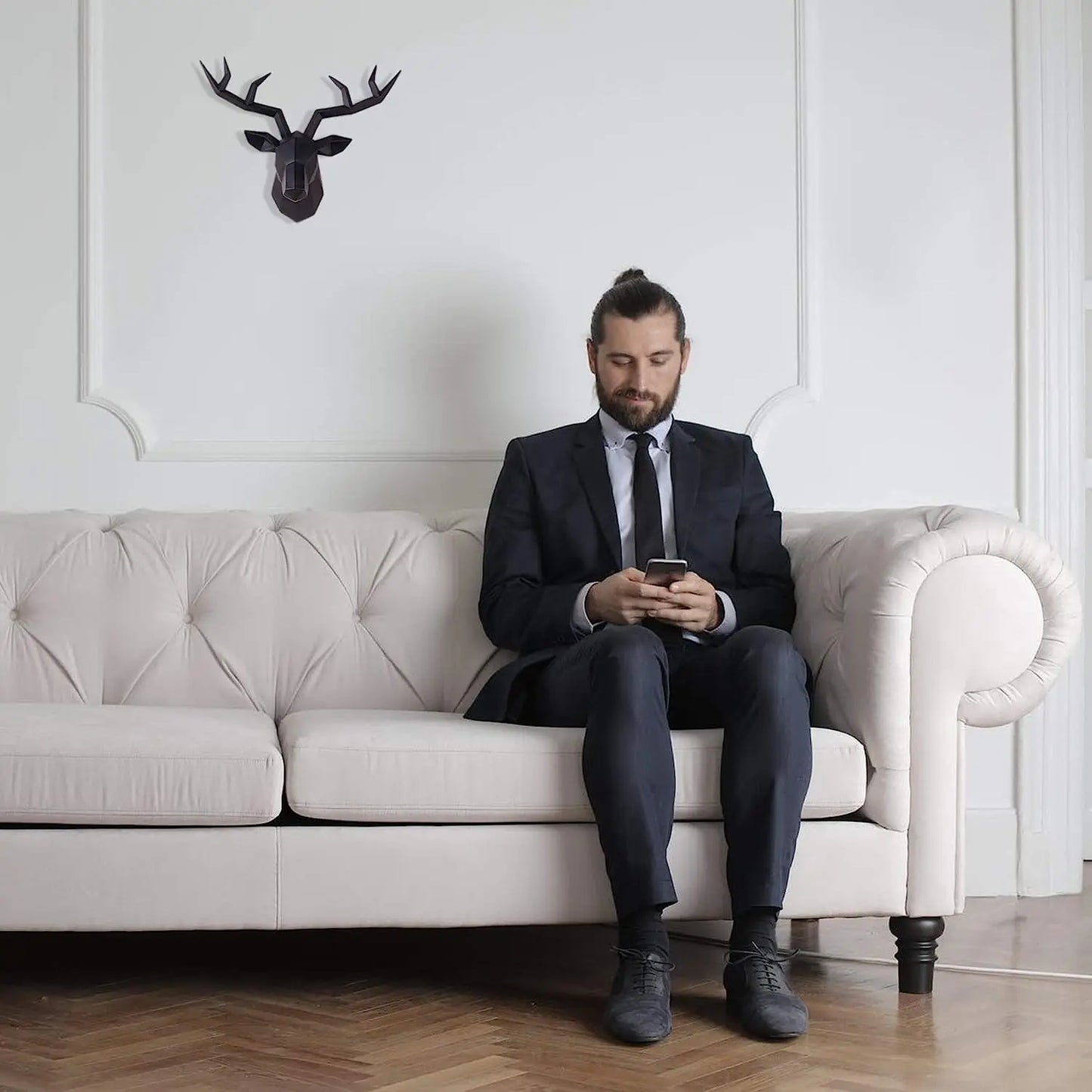 Man in a suit sitting on a white couch using a phone, with a deer head mount on the wall behind him.