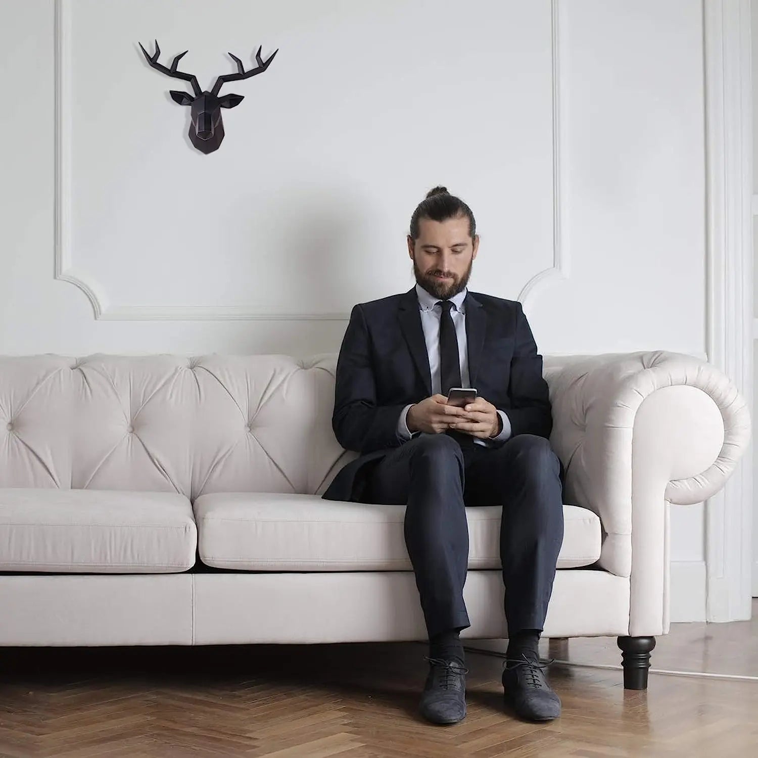Man in a suit sitting on a white couch using a phone, with a deer head mount on the wall behind him.