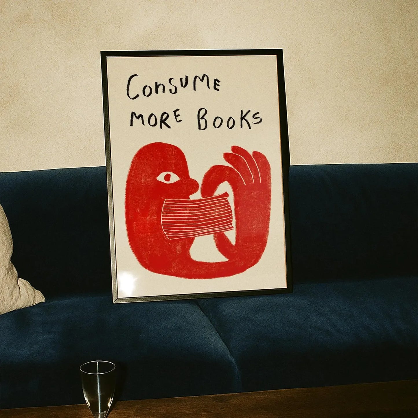 Framed artwork with red illustration and text 'Consume More Books' on a beige wall above a dark blue sofa.