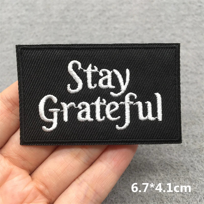 Black patch with 'Stay Grateful' text held in hand against a gray background