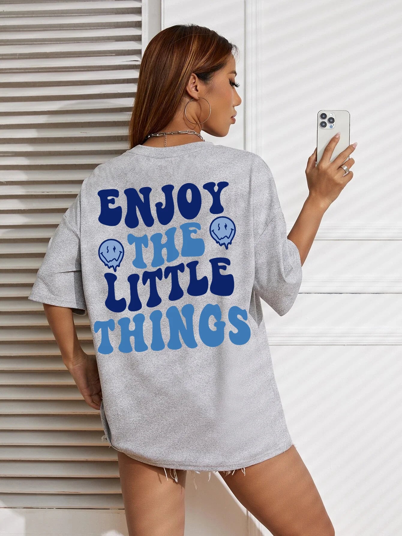 Person wearing a gray t-shirt with 'Enjoy the Little Things' text on the back, standing against a white wall.