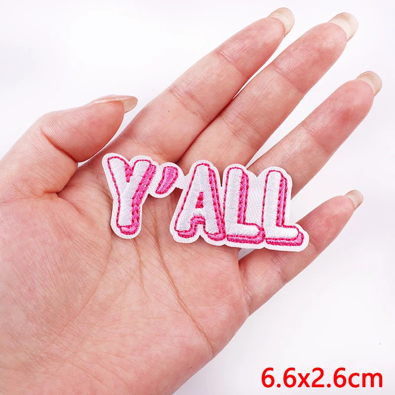 Pink and white 'Y'ALL' patch held in a hand with a white background