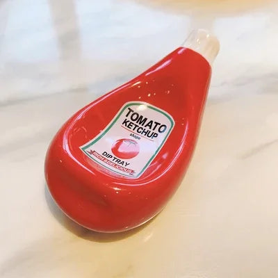 A red ceramic dip tray bottle labeled 'TOMATO KETCHUP' with a white cap, photographed on a light marble-like surface.