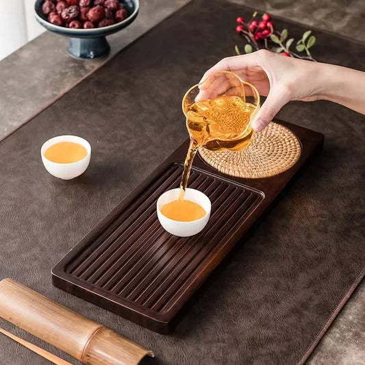 Person pouring tea from a glass teapot into a white cup on a bamboo tea tray.