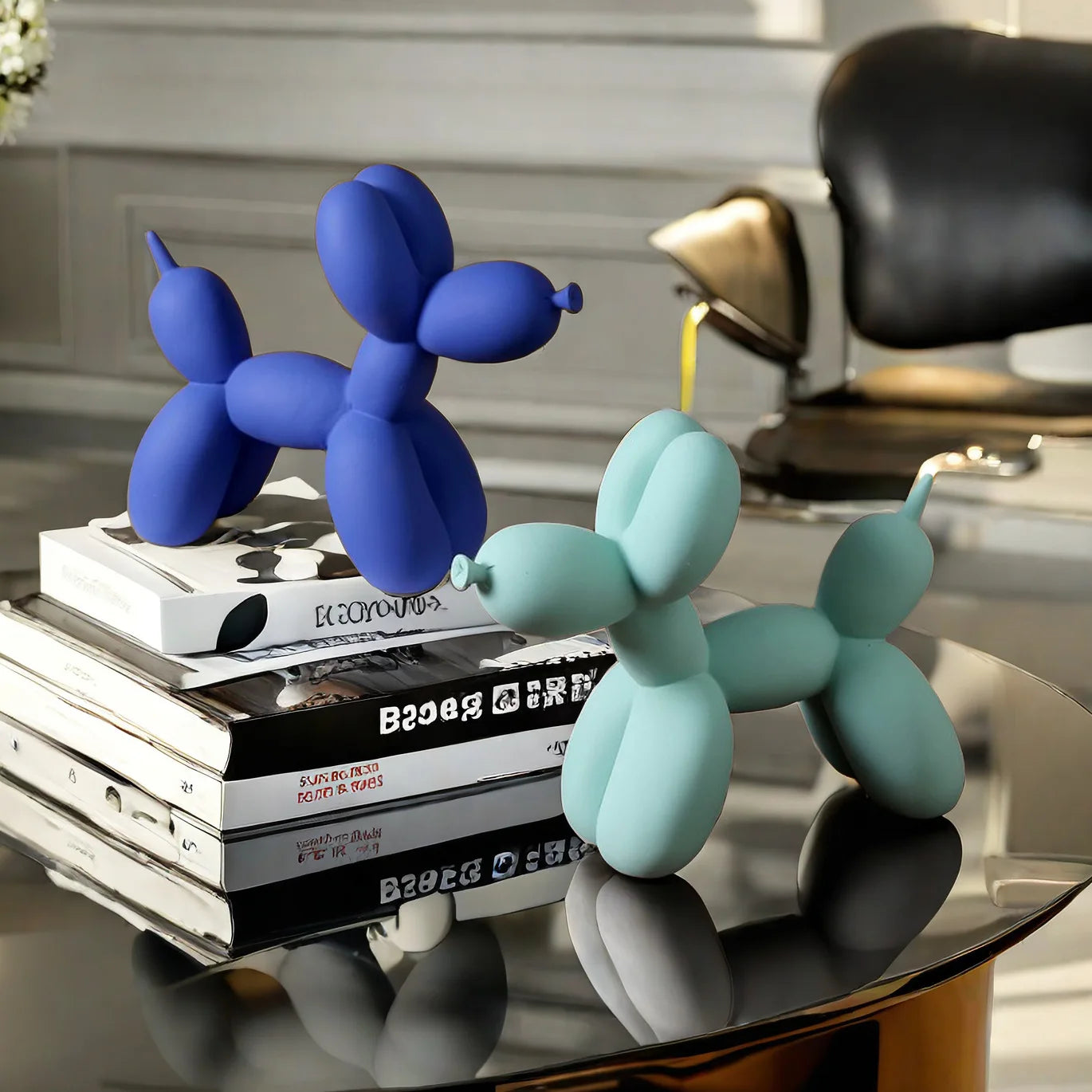 Two balloon dog sculptures, one blue and one green, on a stack of books with a modern interior background.