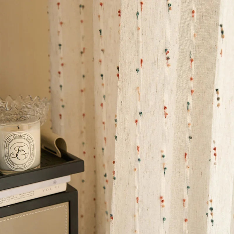 Curtain with small colorful patterns in front of a shelf with a candle and books.