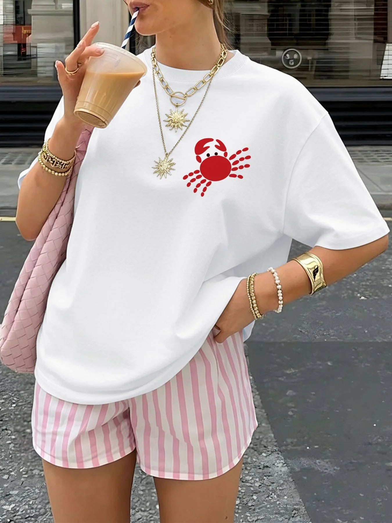 Person wearing a white t-shirt with a red crab design and pink striped shorts, holding a drink.