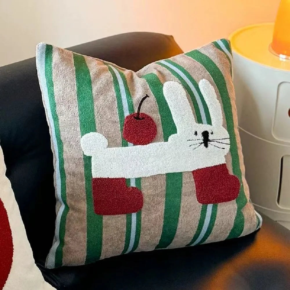 A square throw pillow with a green and beige striped fabric base, featuring a white bunny applique holding a red heart, resting on a dark leather chair.