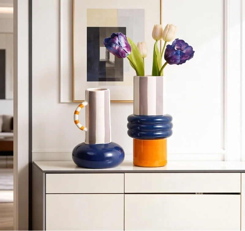 Two modern stacked ceramic vases in a minimalist interior setting. The larger vase on the right features a white upper section, a ribbed navy blue middle section, and an orange base, holding tulips.