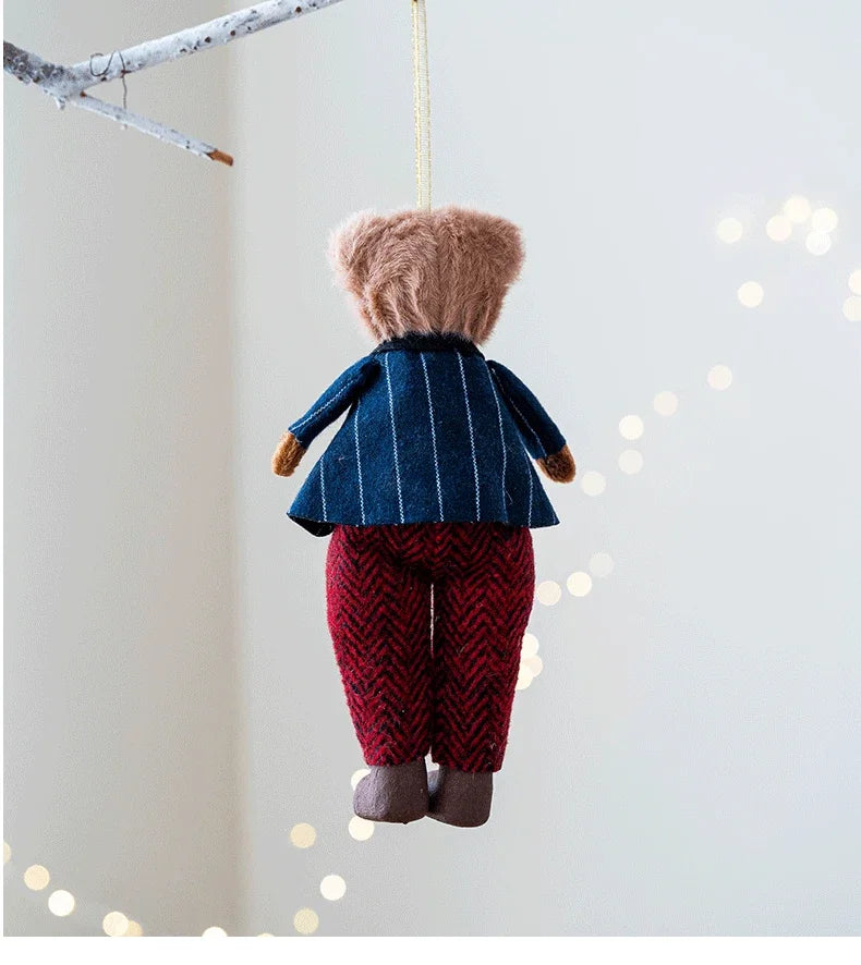 Teddy bear ornament wearing a blue coat and red pants hanging against a light background with blurred lights.