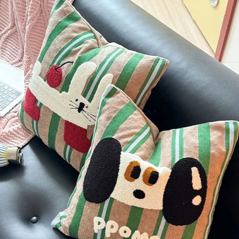 Two decorative throw pillows with green and beige vertical stripes, styled on a black leather sofa. One pillow features a white rabbit holding a red heart, and the other features a dog face with black ears and the text 'PPOM'.