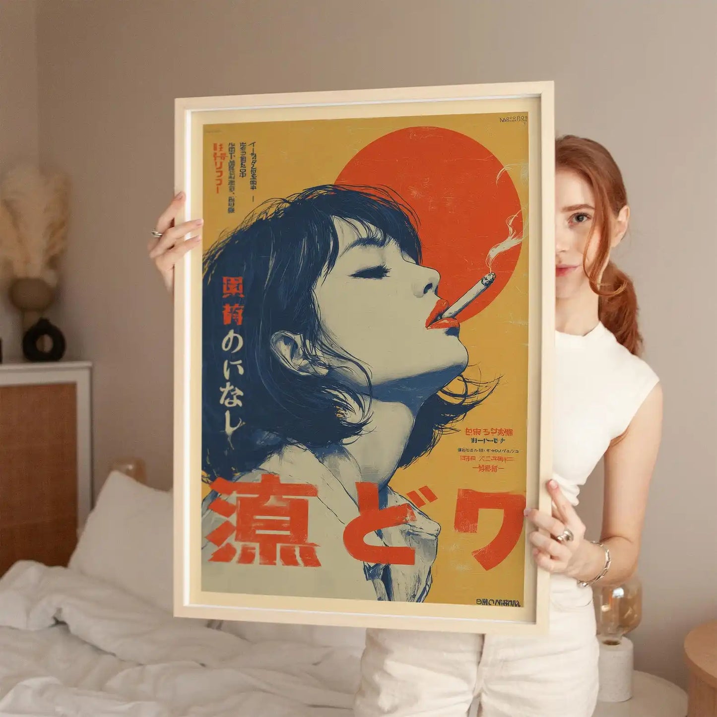 A woman in a white sleeveless top holds up a large framed poster featuring a stylized illustration of a woman with closed eyes, red Japanese text, and a large red sun on a beige background. The room has a neutral beige wall and a bed with white bedding in the background.