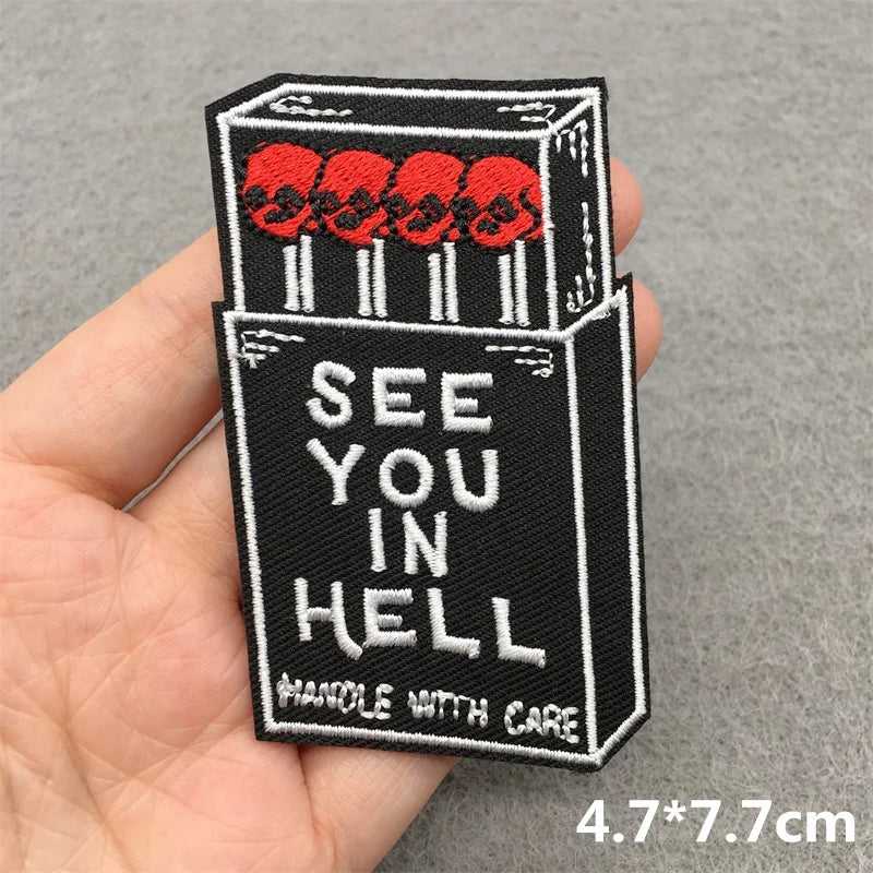 Embroidered patch of a cigarette box with 'See You in Hell' text held in a hand.