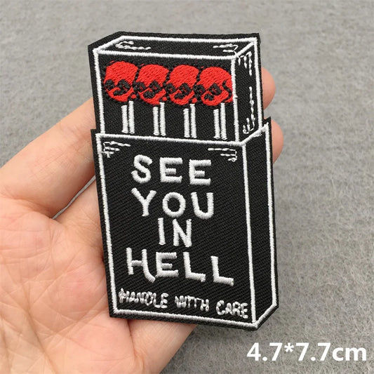 Embroidered patch of a cigarette box with 'See You in Hell' text held in a hand.