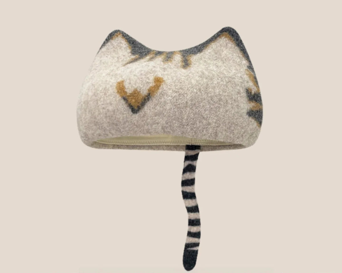 A wool cat-shaped hat in a light beige/gray tone with dark gray and brown tabby-like markings and a black-and-white striped tail, shown on a plain warm beige background
