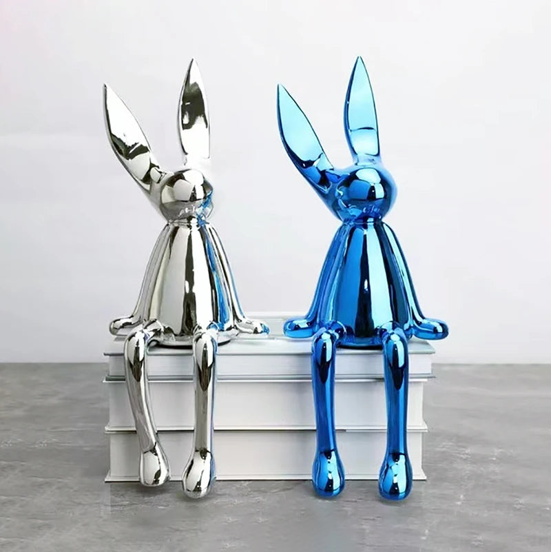 Two metallic rabbit sculptures, one silver and one blue, sitting on a white surface.