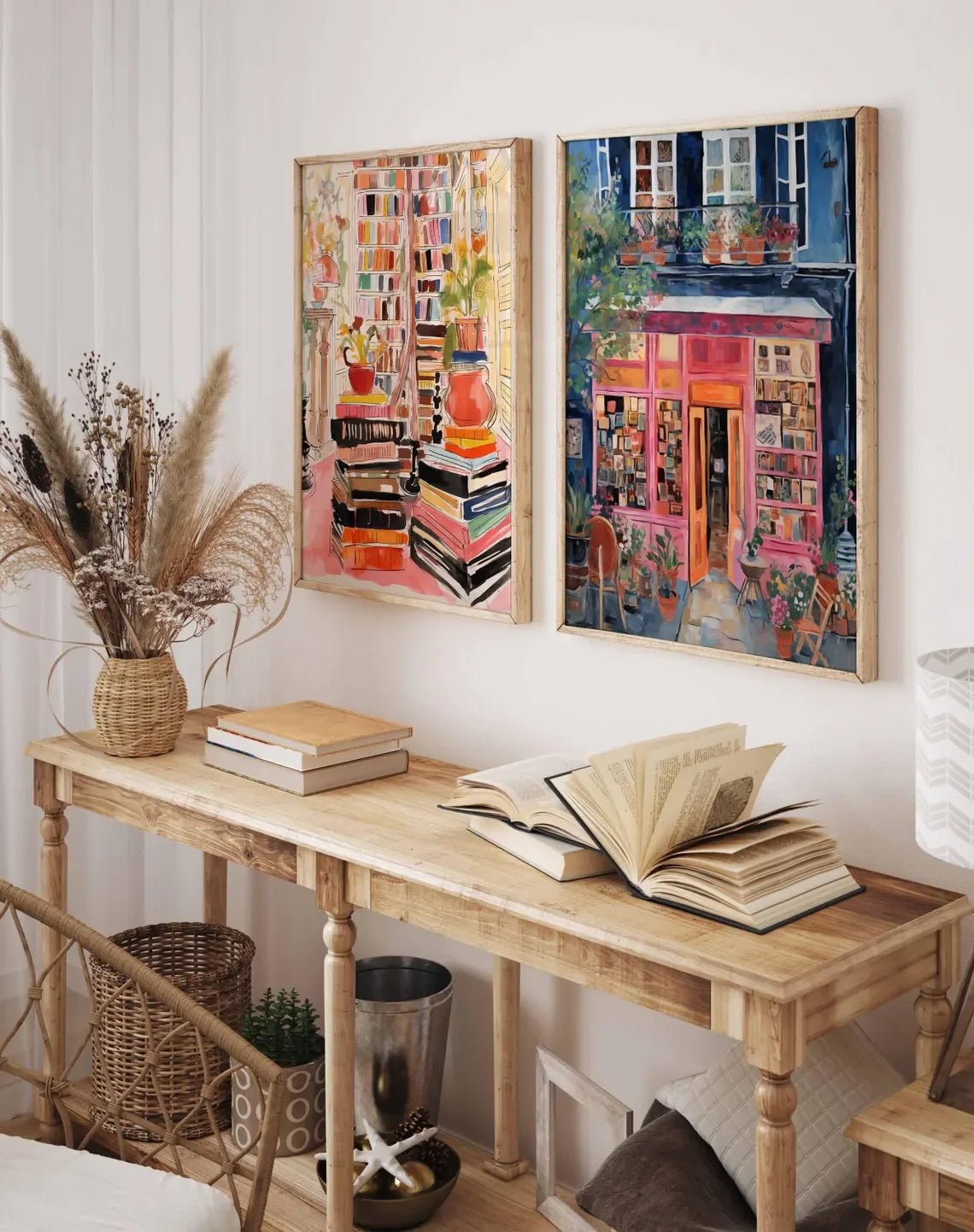 Two framed colorful paintings hanging on a white wall above a wooden console table. The left painting shows stacks of books with warm pink, orange, and cream tones, and the right painting depicts a vibrant storefront scene in pink, blue, and green tones.