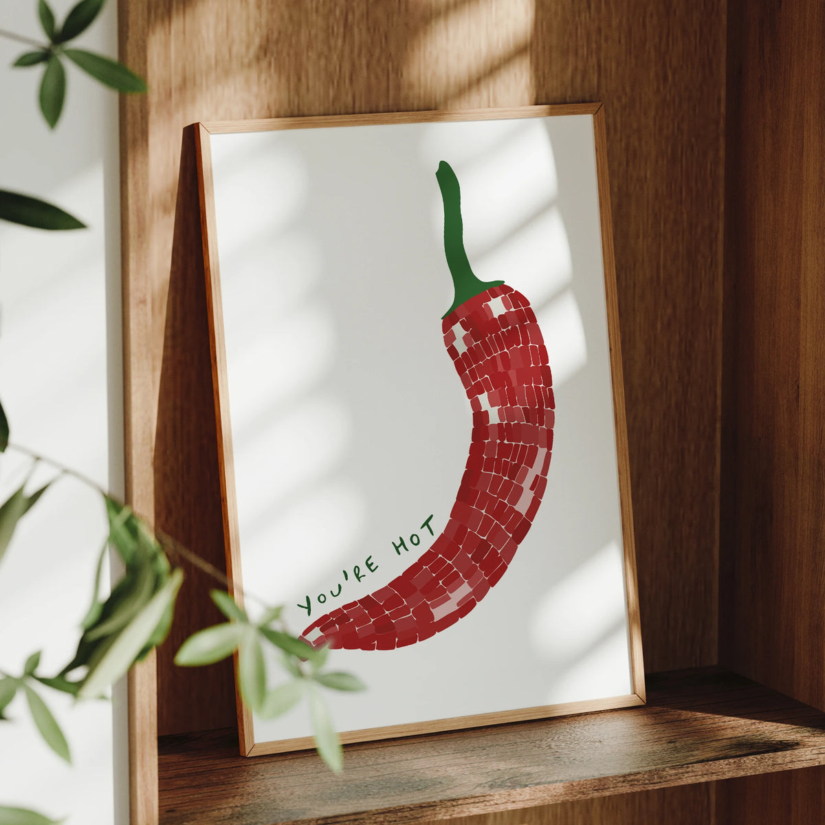 Framed artwork of a red chili pepper with 'You're Hot' text on a wooden shelf.