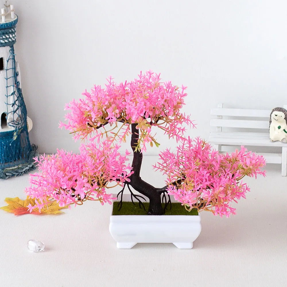 Artificial pink bonsai tree in a white pot on a light gray surface with decorative items in the background.