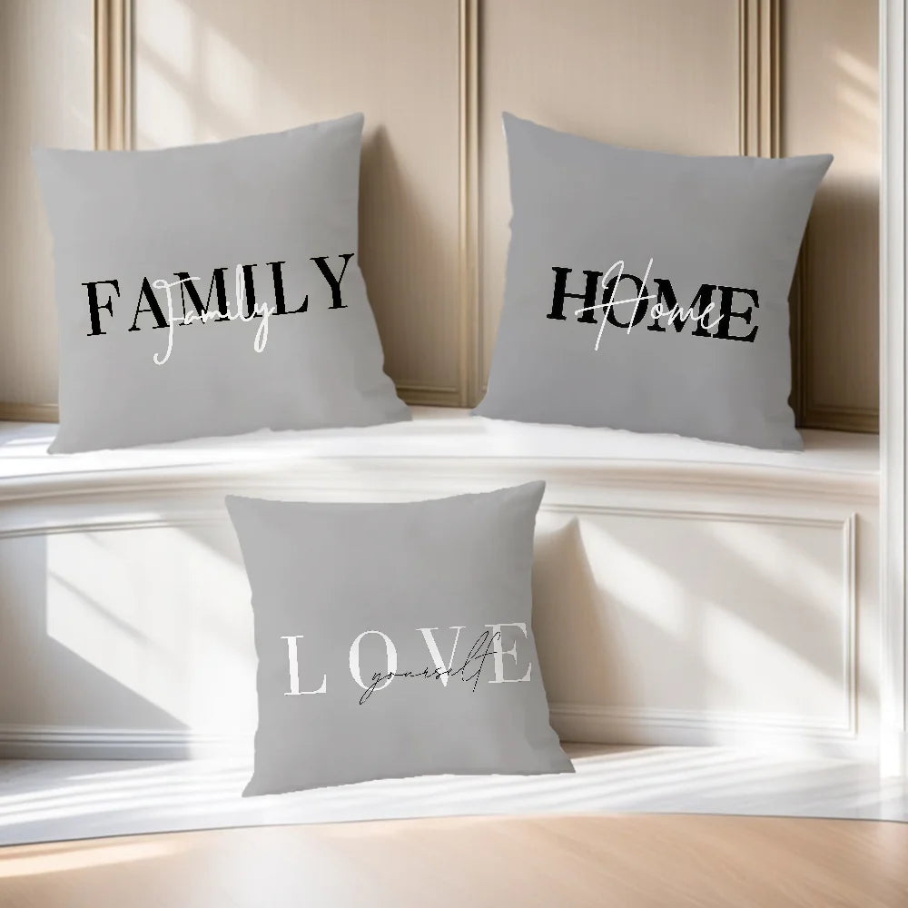 Three gray throw pillows with black and white text reading FAMILY, HOME, and LOVE, arranged on a white bench in a sunlit room.