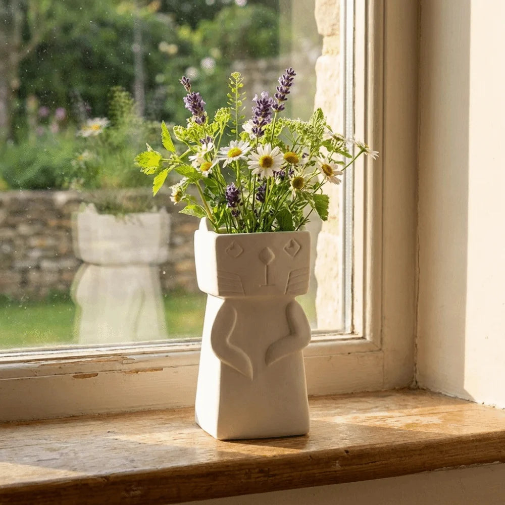 A white ceramic cat-shaped planter holding a bouquet of white daisies and purple lavender, placed on a wooden windowsill with a garden view in the background.