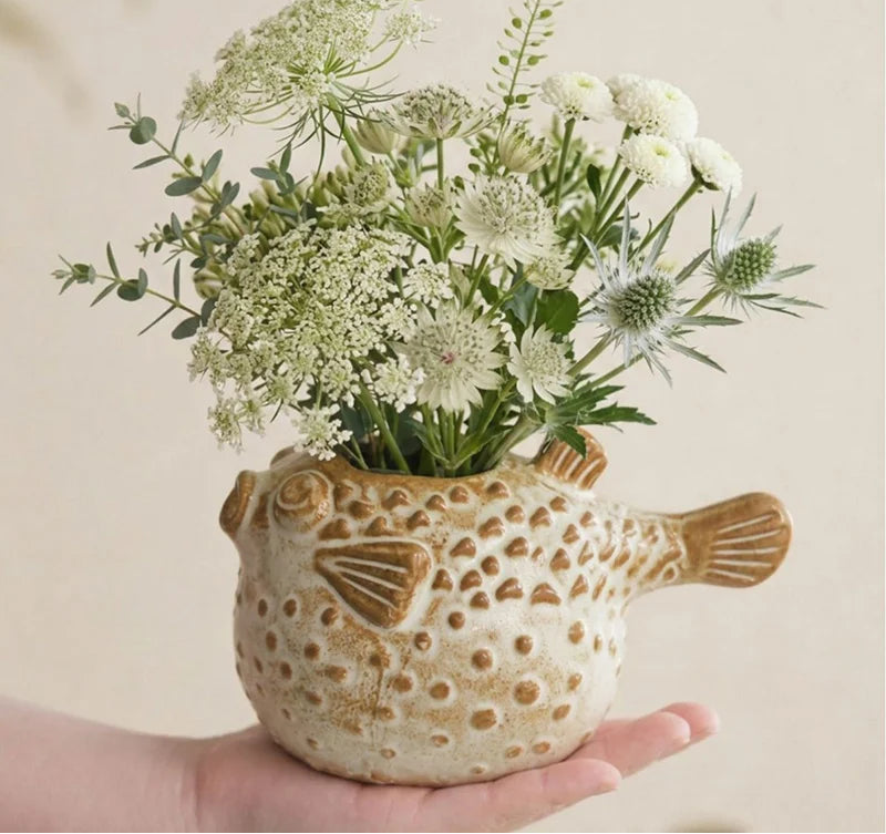 A hand holds a ceramic fish-shaped planter filled with white wildflowers and green foliage, against a plain beige background. The planter is cream-colored with brown speckles and a brown fish tail on the right side.