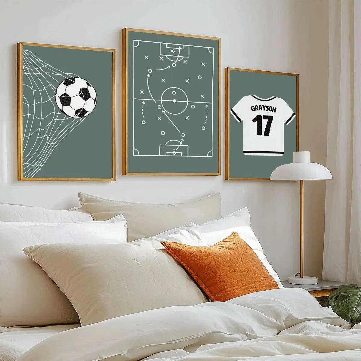 Three framed wall art prints with gold frames hang above a bed in a bedroom. The prints feature a soccer net with a soccer ball, a soccer field strategy diagram, and a white soccer jersey with the name GRAYSON and number 17.