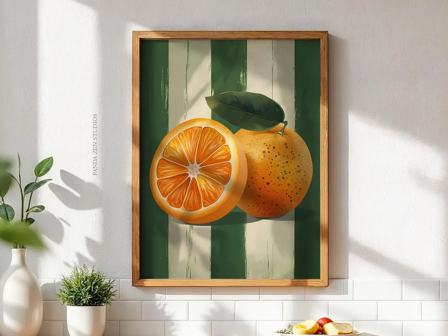 Framed artwork of oranges on a green and white striped background in a bright room.
