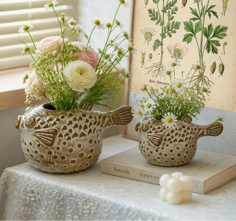 Two ceramic puffer fish planters filled with white and pink flowers, styled on a table with a vintage botanical print and a candle in the background.