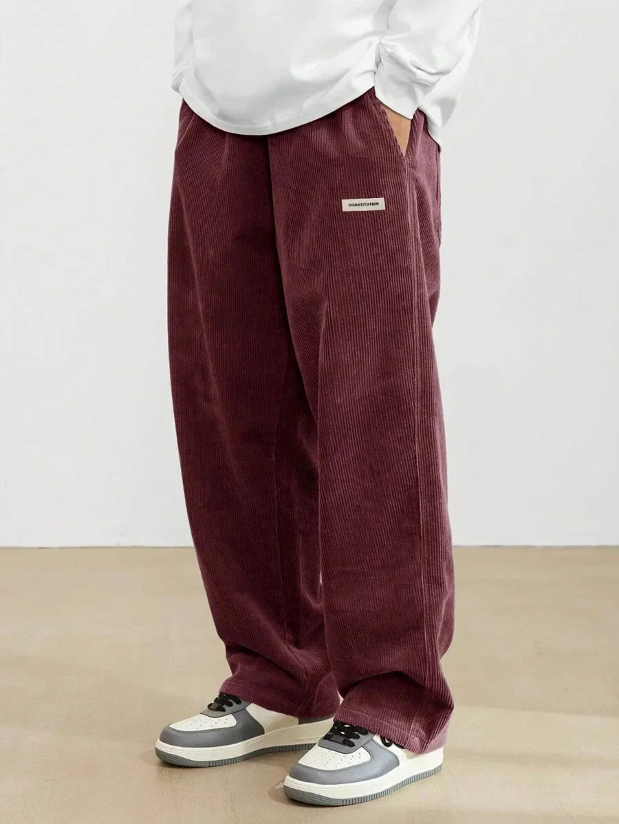 Person wearing maroon corduroy pants with a white shirt and gray sneakers on a plain background