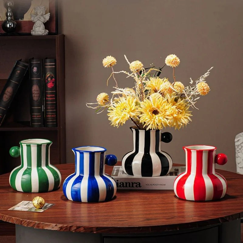 Four decorative ceramic pitchers with bold vertical stripes in green, blue, red, and black-and-white, arranged on a wooden table with yellow flowers in a striped vase, set against a warm brown background.