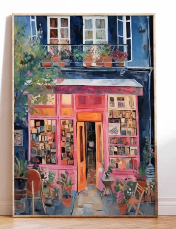 A framed painting of a colorful bookshop storefront with pink and orange doors, blue shutters, and potted plants, leaning against a white wall on a light wood floor.