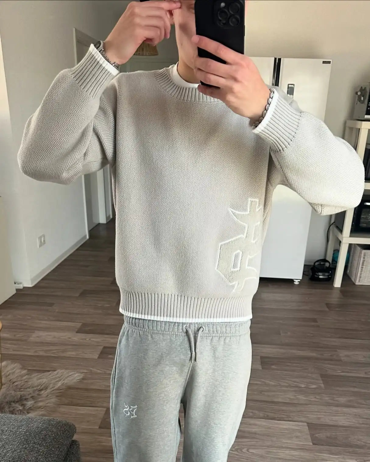 Person wearing a light gray sweater and pants with a brand logo, taking a mirror selfie.
