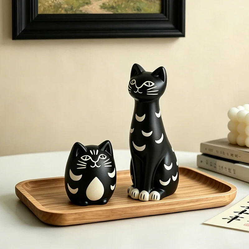Two black cat figurines with white crescent moon patterns, one small and one large, displayed on a wooden tray on a tabletop.