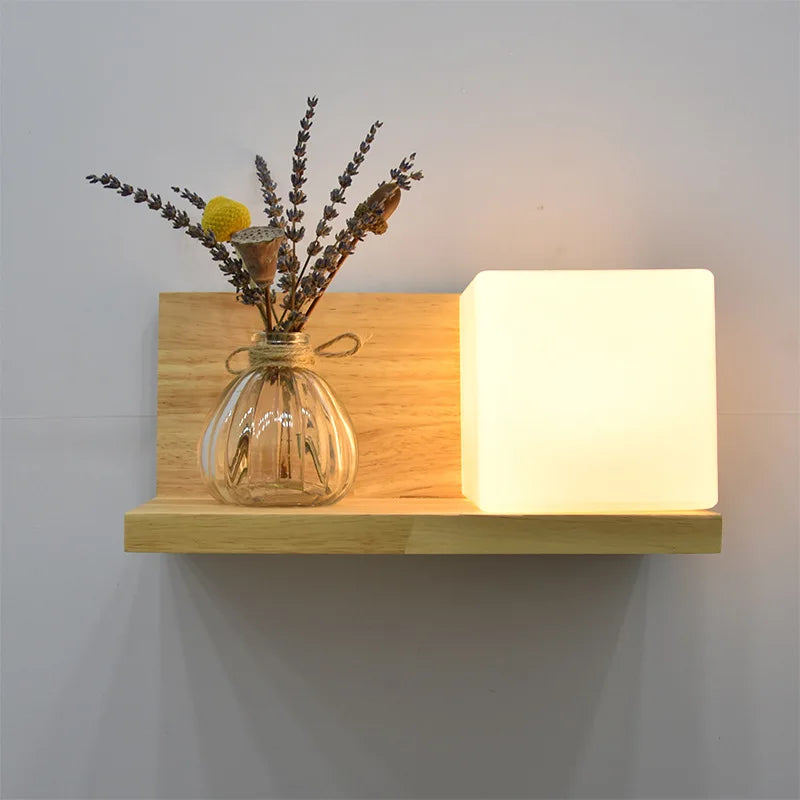 Wooden shelf with a glass vase containing dried flowers and a square light fixture on a plain wall.