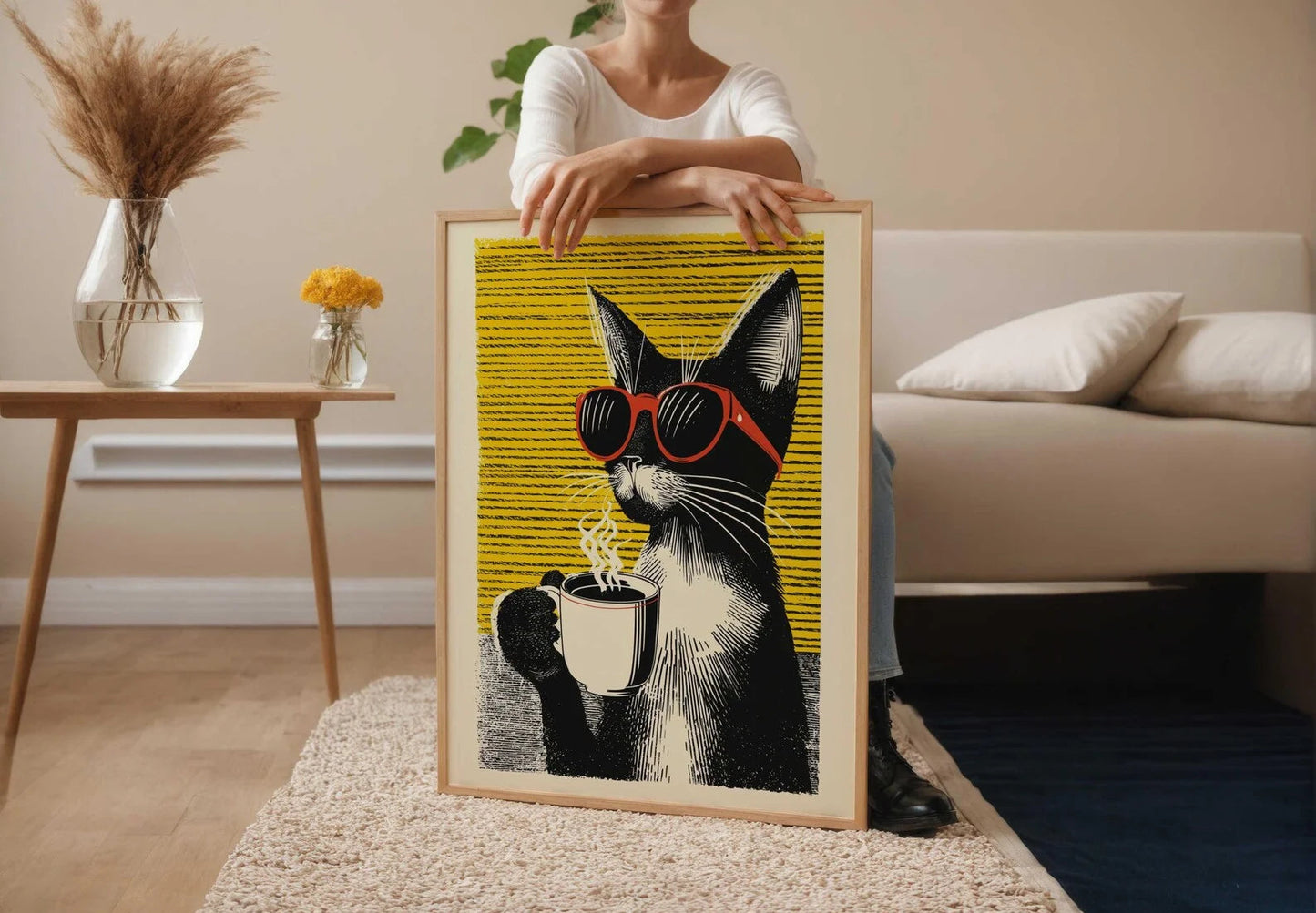 Person holding a framed artwork of a cat wearing sunglasses and a coffee cup in a living room.