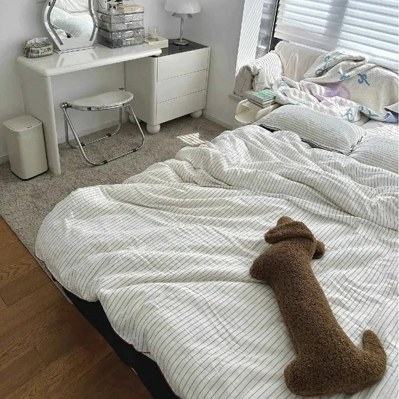 Bedroom with a bed featuring a checkered duvet and a brown plush dachshund-shaped cushion, next to a desk and chair.