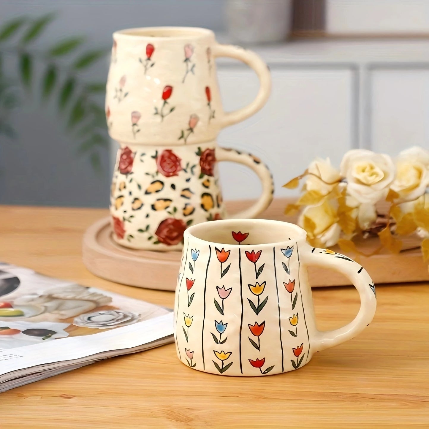 Set of floral-patterned mugs on a wooden surface with a magazine and flowers in the background.