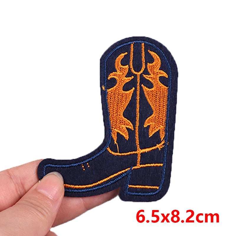 Embroidered patch of a cowboy boot with orange design on a blue background, held by a hand with measurement text '6.5x8.2cm'.