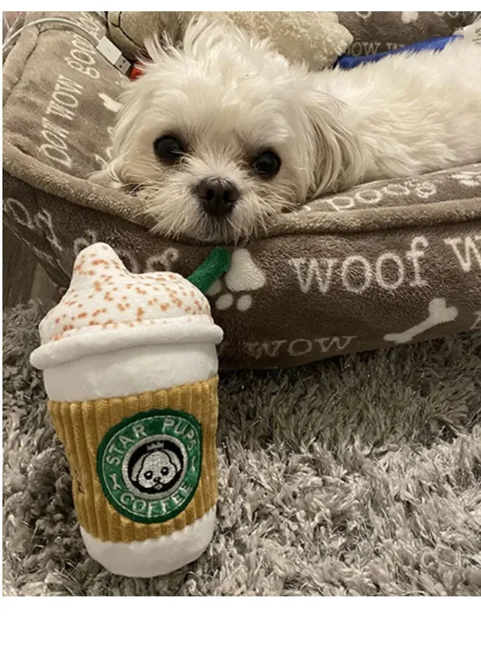 Dog toy shaped like a Starbucks coffee cup with a small white dog lying on a cushion in the background.